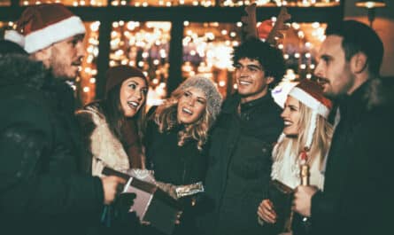 Sechs Kolleg:innen mit Weihnachtsmützen und Geschenken lachen vor warmen Lichterketten – stimmungsvolle Szene einer Firmen‑Weihnachtsfeier.