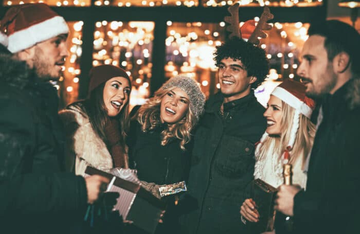 Sechs Kolleg:innen mit Weihnachtsmützen und Geschenken lachen vor warmen Lichterketten – stimmungsvolle Szene einer Firmen‑Weihnachtsfeier.